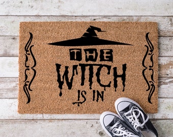 May include: A brown coir doormat with the text "The Witch is in" in black lettering. The text is stylized with dripping letters and a witch's hat above the word "The". The doormat has decorative scrollwork on the sides.