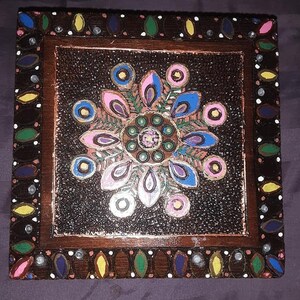 May include: A wooden box with a painted mandala design. The mandala is in shades of pink, blue, green, and gold. The box is surrounded by a brown frame with a decorative border.