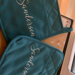 Puede incluir: Dos agarraderas de color turquesa con el nombre "Sanderson" en escritura blanca. Las agarraderas tienen un patrón de diamantes acolchados y un lazo para colgar. Fabricadas con tela duradera.