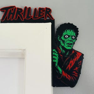Peut inclure: Décoration de porte noire et rouge avec le mot "Thriller" en lettres rouges. Une figurine de zombie à la peau verte et à la veste rouge regarde autour du cadre de la porte. Thème d'Halloween ou d'horreur.