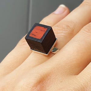 Puede incluir: Un anillo cuadrado de madera con una incrustación roja. El anillo está hecho de madera oscura y tiene una banda plateada.
