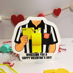 May include: Valentine's Day card shaped like a referee's shirt, with black and white stripes. The card features a yellow whistle and the text "Whistling You a Happy Valentine's Day!"