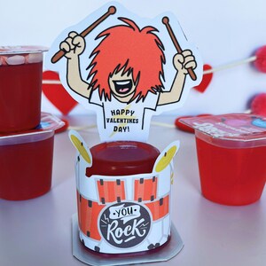 May include: Red gelatin cups with a Valentine's Day theme. A cartoon drummer with red hair and drumsticks is featured on a decorative drum-shaped container. The text "Happy Valentine's Day!" is on the drummer's shirt, and "You Rock" is on the drum.