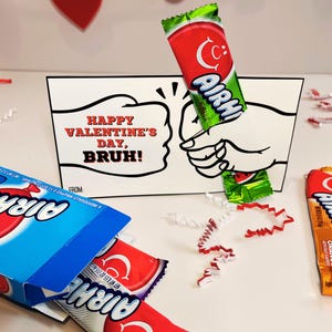 Op de afbeelding: Een Valentijnsdagkaart met de tekst "Happy Valentine's Day, Bruh!" toont een cartoonillustratie van twee handen die elkaar aanraken. Airheads snoep en een doos Airheads staan ook op de afbeelding.