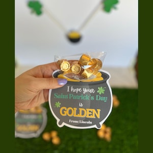 Puede incluir: Una olla de oro negra y gris con un trébol verde y el texto "Espero que tu día de San Patricio sea GOLDEN. De: Lincoln." La olla está llena de monedas de chocolate envueltas en papel dorado.