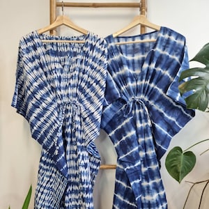May include: Two blue and white tie-dye kaftans hanging on wooden hangers. The kaftans have a V-neck and are made of a lightweight fabric.