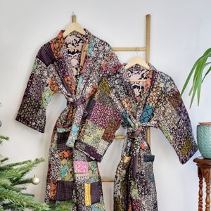 Puede incluir: Dos batas acolchadas de patchwork colgadas de una escalera de madera. Las batas son en tonos de negro, marrón, rosa, azul y verde. Las batas tienen un cinturón de atar y mangas largas.