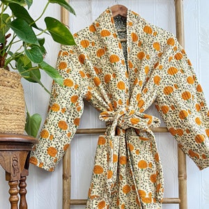 Op de afbeelding: Een lange, witte badjas met een bloemenpatroon in tinten oranje en groen. De badjas heeft een strikceintuur en een shawlkraag. Hij hangt aan een houten ladder.