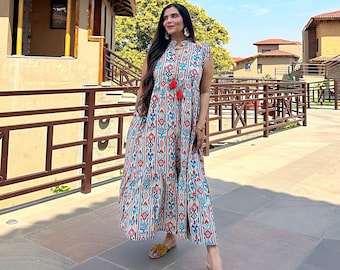 May include: A woman wearing a long, flowing, white dress with a red, blue, and white geometric pattern. The dress has a tassel detail at the neckline. She is standing on a paved walkway in front of a building with a terracotta roof.