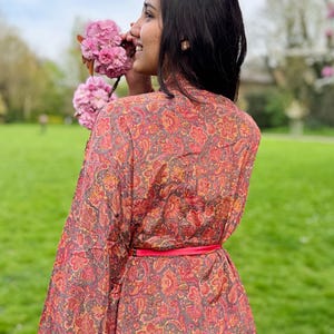 Può includere: Una donna indossa un lungo kimono rosso e oro con motivo paisley e una cintura rossa. È in piedi in un parco e annusa dei fiori rosa.