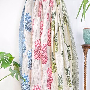 May include: Three kantha quilts with a white background and pineapple print in blue, pink, and green. The quilts are folded and draped over a wooden rod.