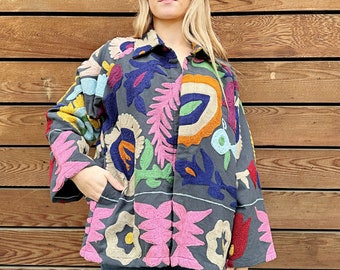 May include: A dark gray jacket with colorful floral embroidery. The jacket features a button-up front, long sleeves, and pockets. The embroidery includes pink, blue, orange, green, and red floral designs. The model is wearing blue jeans.
