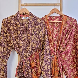 Op de afbeelding: Twee lange, gebloemde badjassen. De badjas links is donkerpaars met een gouden bloemenpatroon. De badjas rechts is bordeauxrood met een gouden bloemenpatroon. Beide badjassen hebben een strik in de taille.