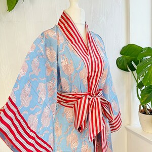 May include: A light blue kimono-style robe with a repeating pattern of orange tigers. The robe features red and white striped trim on the collar, cuffs, and belt. The belt is tied in a bow at the waist, creating a stylish and comfortable look.