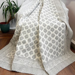 Könnte beinhalten: Eine weiße Baumwolldecke mit einem grauen Blattmuster. Die Decke ist gefaltet und über einen Stuhl gelegt.