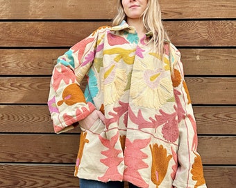 Op de afbeelding: Een beige geborduurde jas met een bloemenpatroon in tinten roze, oranje, geel en blauw. De jas heeft een kraag en lange mouwen.