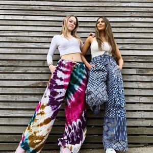 May include: Two people wearing colourful pants. One person is wearing a white crop top and tie-dye pants with shades of purple, pink, green, and brown. The other person is wearing a white tank top and blue patterned pants. Both people are standing in front of a wooden wall.