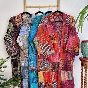 Op de afbeelding: Drie lange, patchwork gequilte gewaden in tinten van bruin, teal en rood hangen aan een houten ladder. De gewaden zijn gemaakt van verschillende stoffen en patronen, en ze hebben een ontspannen, bohemian stijl.