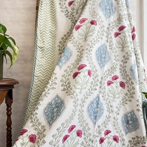 May include: White fabric with a floral pattern featuring pink flowers, blue diamond shapes, and green leaves. The fabric is draped over a wooden rod, revealing a green and white chevron pattern on the reverse side.