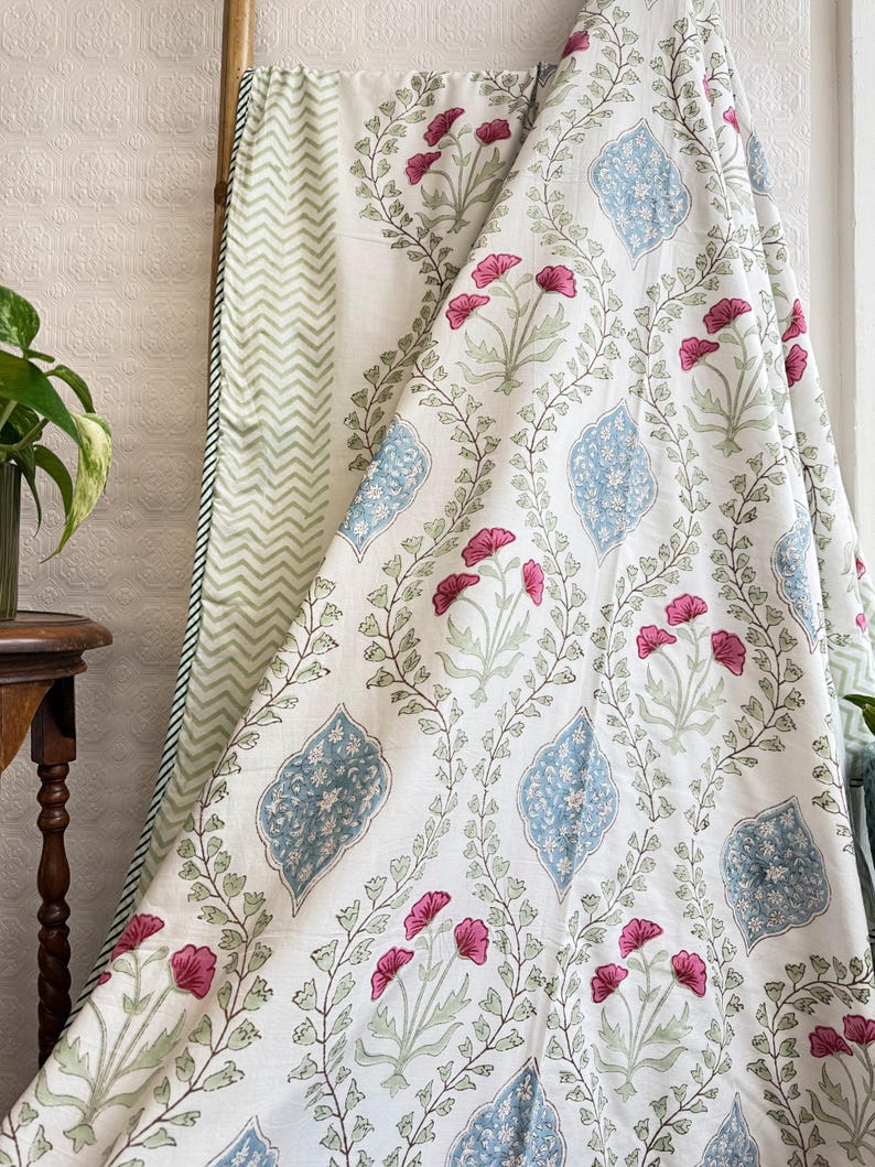 May include: White fabric with a floral pattern featuring pink flowers, blue diamond shapes, and green leaves. The fabric is draped over a wooden rod, revealing a green and white chevron pattern on the reverse side.