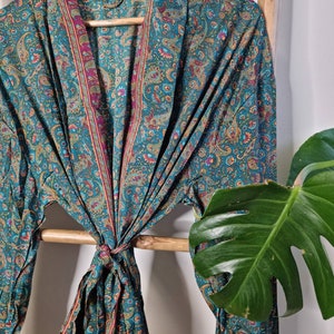 May include: A long-sleeved robe featuring a teal base color with an intricate paisley pattern in shades of magenta, gold, and orange. The garment has a wide shawl collar and a matching fabric tie belt at the waist, creating a draped silhouette.
