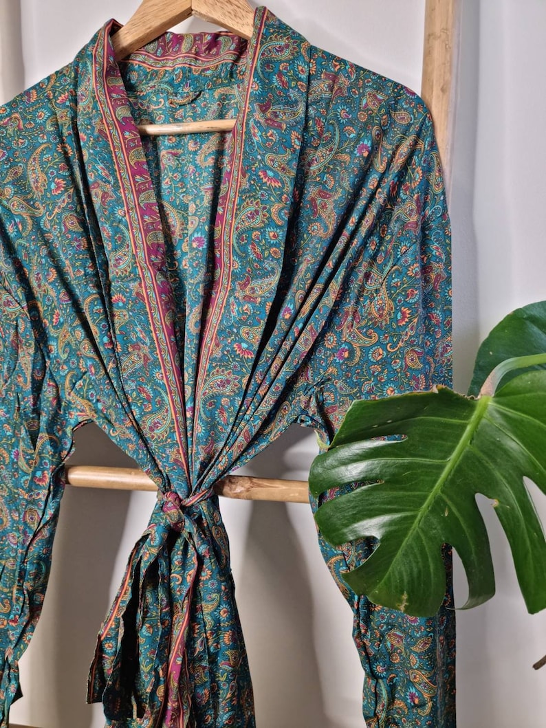 May include: A long-sleeved robe featuring a teal base color with an intricate paisley pattern in shades of magenta, gold, and orange. The garment has a wide shawl collar and a matching fabric tie belt at the waist, creating a draped silhouette.