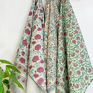 May include: Three floral patterned textiles hanging from a wooden rack. The textiles feature a variety of colours, including red, green, and cream. The floral patterns are detailed and intricate, creating a visually appealing display.