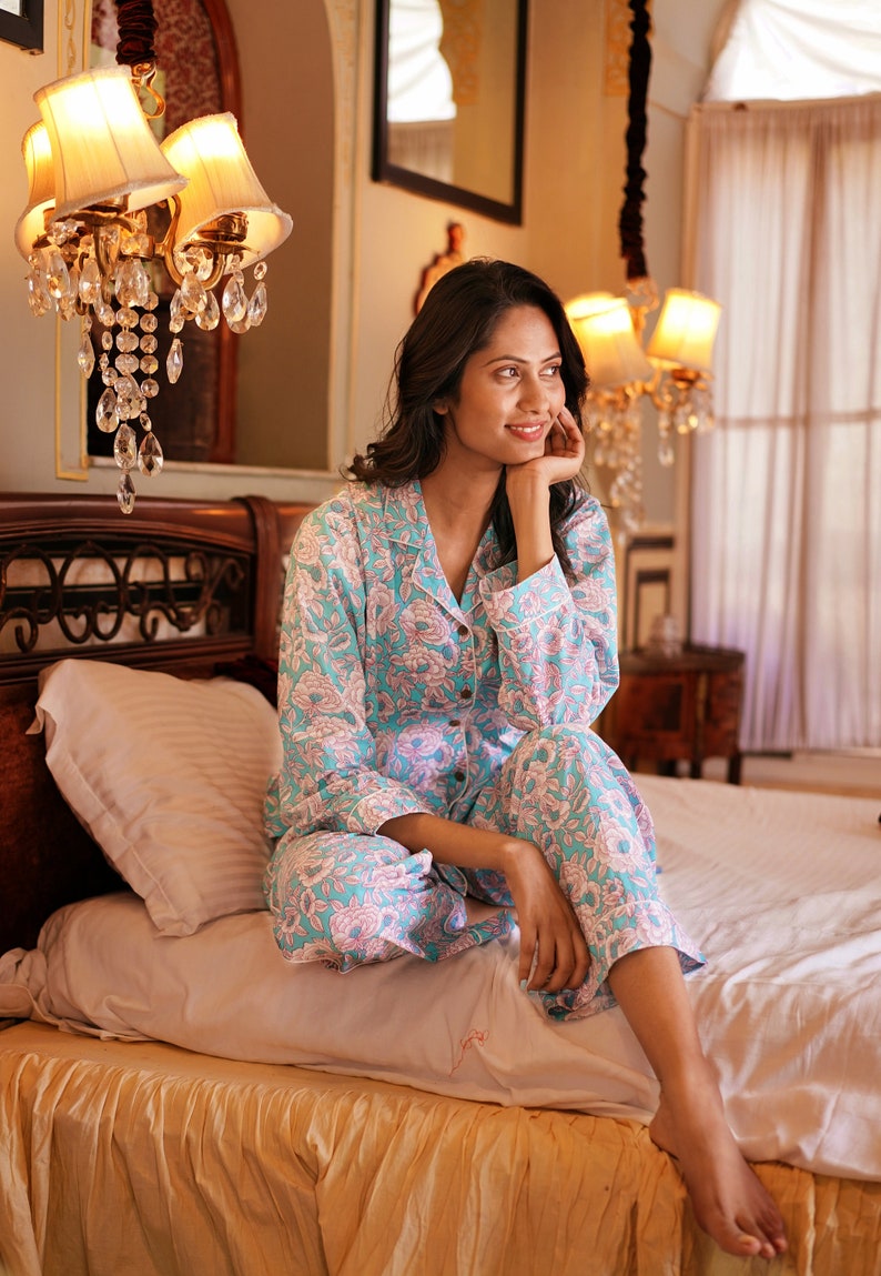 May include: A woman wearing a light blue and pink floral print pyjama set sits on a bed with a white bedspread and a gold bed skirt.