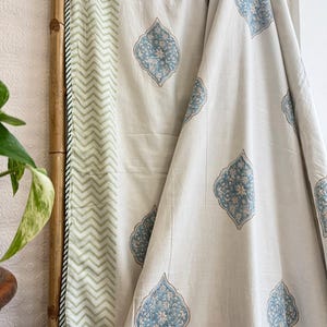 May include: Off-white fabric with a repeating blue floral pattern. A section of the fabric features a green and white chevron design. The fabric is draped over a bamboo pole, with a plant in the foreground.