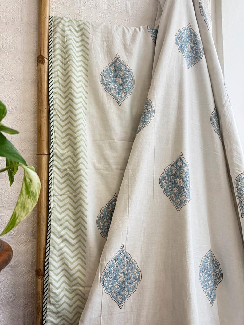 May include: Off-white fabric with a repeating blue floral pattern. A section of the fabric features a green and white chevron design. The fabric is draped over a bamboo pole, with a plant in the foreground.
