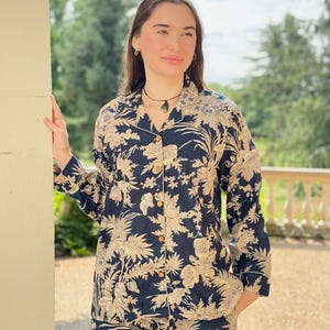 Puede incluir: Conjunto de pijama azul marino con un estampado floral beige. El conjunto incluye una camisa con botones y pantalones a juego. La camisa tiene cuello y botones de madera. La persona lleva un collar y pendientes.