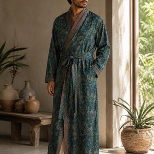 May include: A floor-length teal robe featuring a detailed paisley pattern in shades of orange, pink, and green. The garment includes a shawl collar with a contrasting border, long sleeves, and a matching fabric tie belt at the waist.