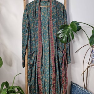 May include: A long-sleeved duster robe in a deep teal color featuring an intricate paisley pattern in shades of gold, pink, and blue. The garment has an open front design with a contrasting vertical border trim and two large patch pockets.