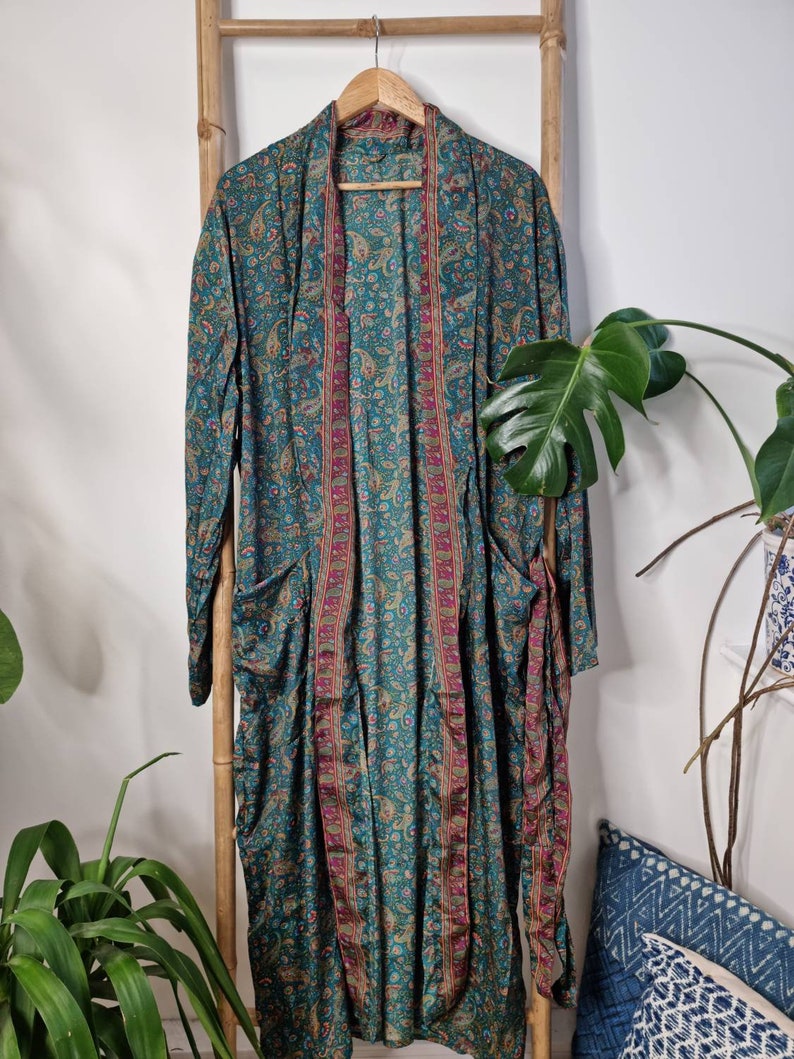 May include: A long-sleeved duster robe in a deep teal color featuring an intricate paisley pattern in shades of gold, pink, and blue. The garment has an open front design with a contrasting vertical border trim and two large patch pockets.