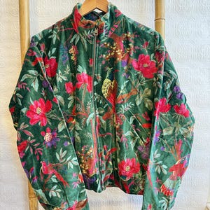 May include: A green velvet jacket with a vibrant floral and bird pattern. The jacket features a high collar, a full-length zipper, and elastic cuffs. The design includes red flowers, birds, and foliage.