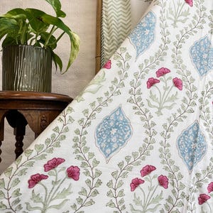 May include: White fabric with a floral pattern featuring pink flowers, blue leaf-shaped designs, and green vines. A potted plant and a wooden frame are visible in the background. The fabric is likely used for home decor or textiles.