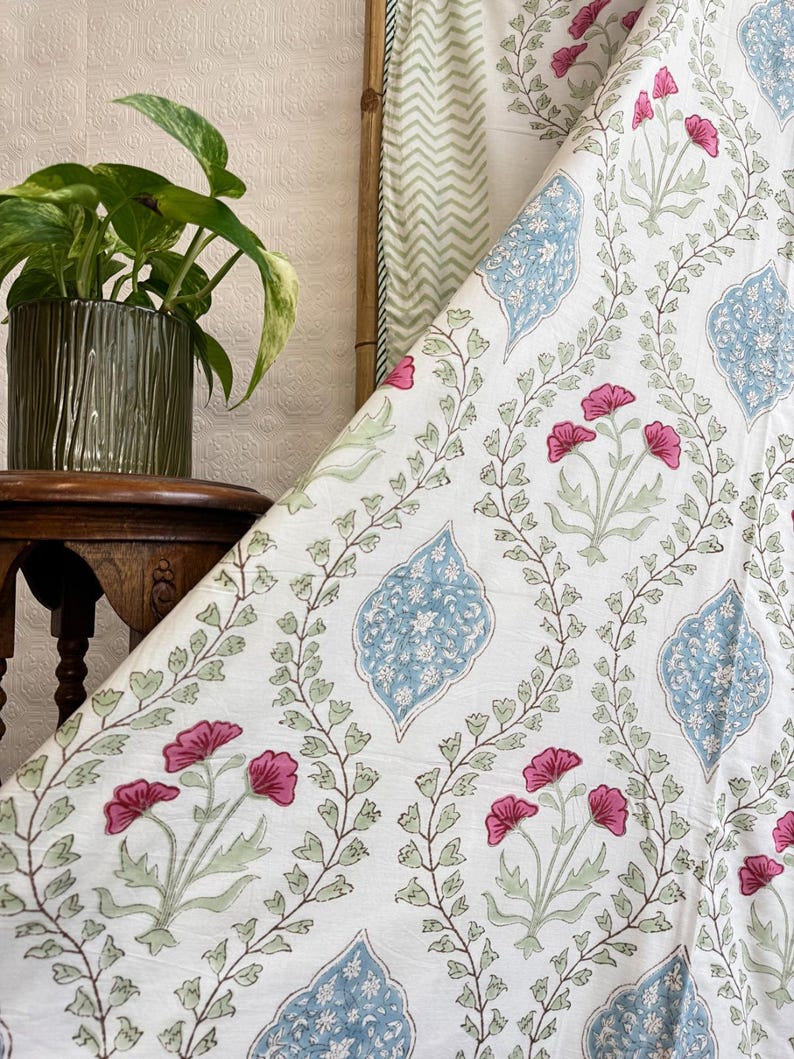 May include: White fabric with a floral pattern featuring pink flowers, blue leaf-shaped designs, and green vines. A potted plant and a wooden frame are visible in the background. The fabric is likely used for home decor or textiles.