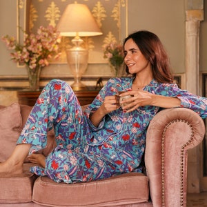 Peut inclure: Une femme portant un pyjama bleu et rose &agrave; imprim&eacute; floral est assise sur un canap&eacute; en velours rose. Elle tient une tasse dans sa main et regarde vers la droite.