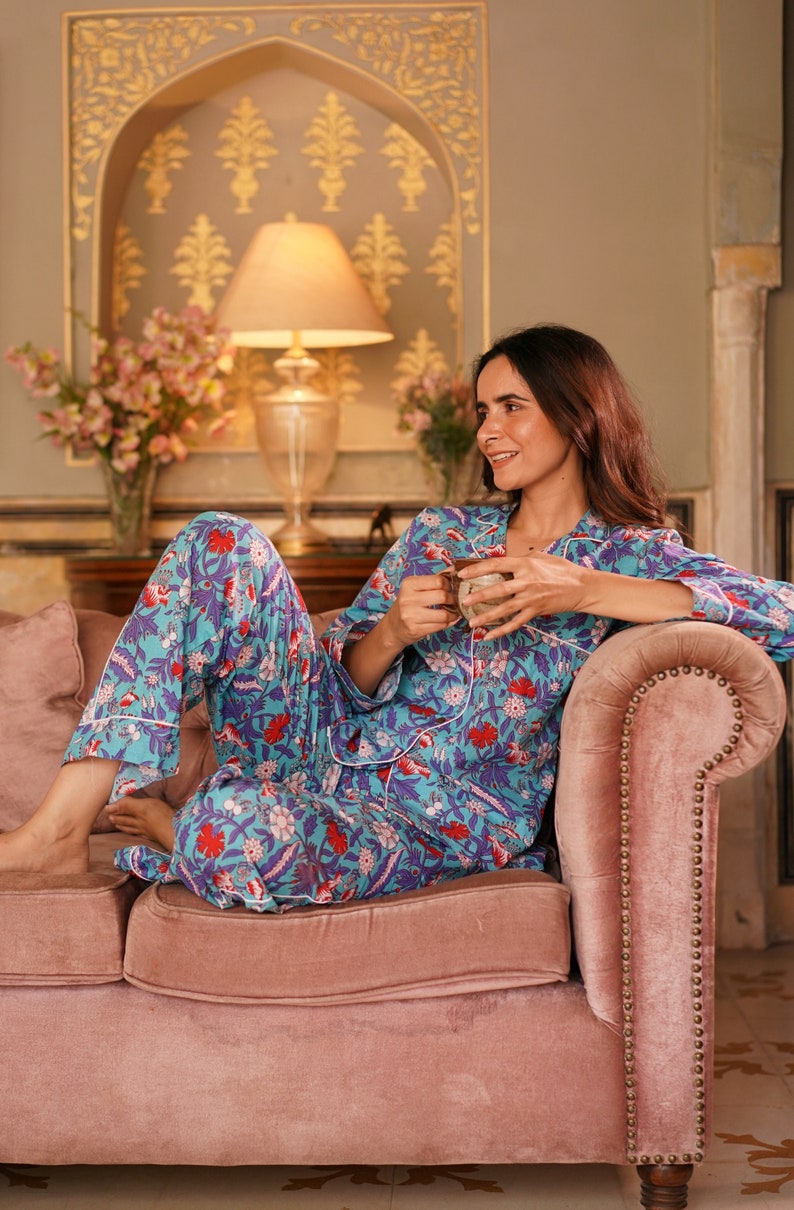 Peut inclure: Une femme portant un pyjama bleu et rose &agrave; imprim&eacute; floral est assise sur un canap&eacute; en velours rose. Elle tient une tasse dans sa main et regarde vers la droite.