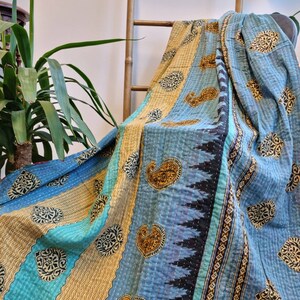 May include: A blue and gold kantha quilt with paisley and floral patterns. The quilt is folded and draped over a wooden ladder.