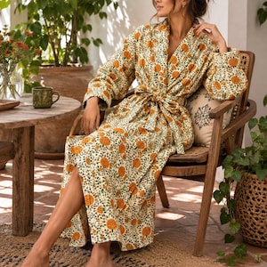 May include: A long-sleeved, floor-length quilted robe featuring a cream-coloured base with a repeating pattern of orange marigold flowers and green foliage. The robe includes a matching fabric tie belt at the waist and a wide shawl collar.