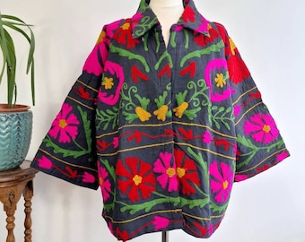 Op de afbeelding: Een donkergrijze jas met een bloemenborduurpatroon in tinten roze, rood, geel en groen. De jas heeft een kraag en een knoopsluiting.