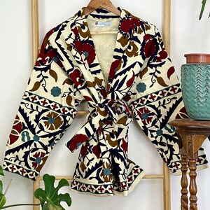 Könnte beinhalten: Ein cremefarbener Kimono mit einem floralen Muster in Rot-, Blau-, Braun- und Grüntönen. Der Kimono hat einen Bindegürtel und lange Ärmel.