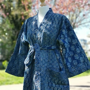 Könnte beinhalten: Eine blaue Baumwollrobe mit einem weißen Stickmuster. Die Robe hat einen Bindegürtel und eine Tasche auf der linken Seite.