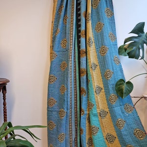 May include: A blue and teal kantha quilt with a paisley pattern and gold accents. The quilt is folded and draped over a wooden rack.