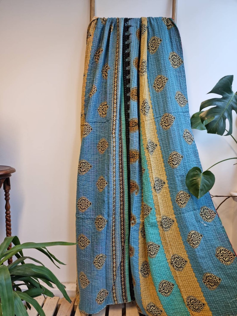 May include: A blue and teal kantha quilt with a paisley pattern and gold accents. The quilt is folded and draped over a wooden rack.