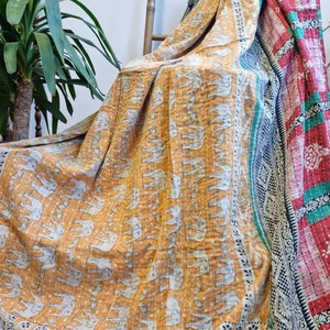 May include: A kantha quilt with a mustard yellow background and a pattern of white elephants. The quilt has a border with a floral design in shades of green, black, and white. The quilt is folded and draped over a wooden chair.