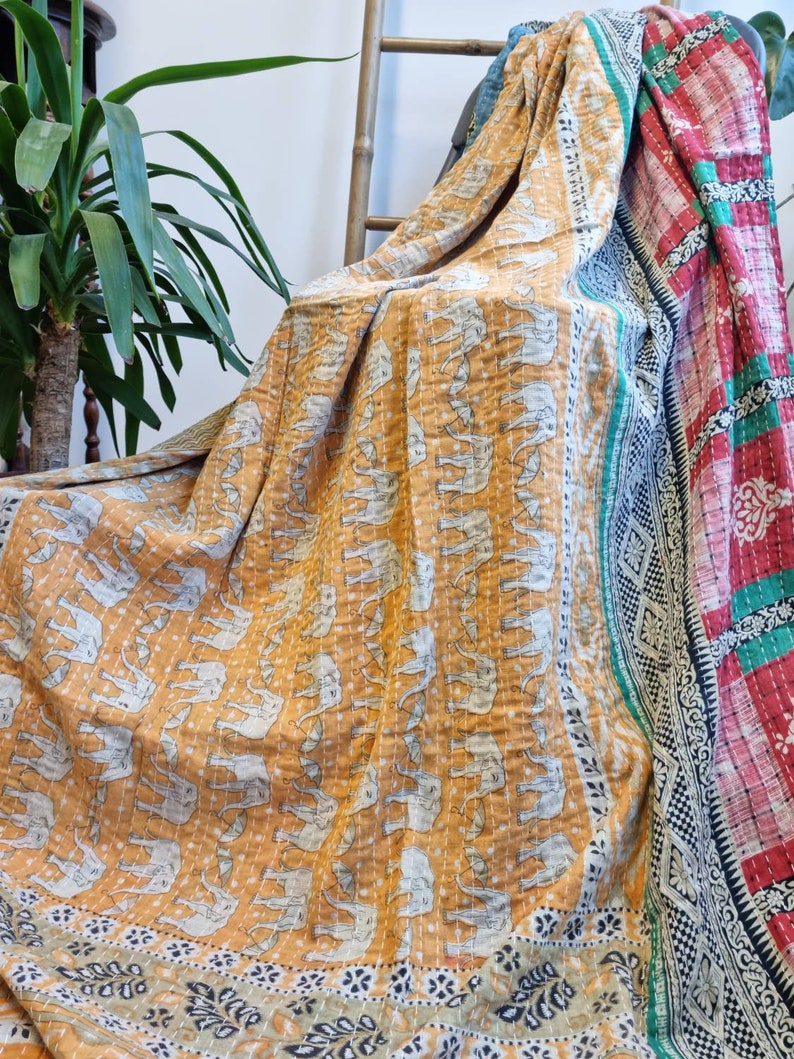 May include: A kantha quilt with a mustard yellow background and a pattern of white elephants. The quilt has a border with a floral design in shades of green, black, and white. The quilt is folded and draped over a wooden chair.