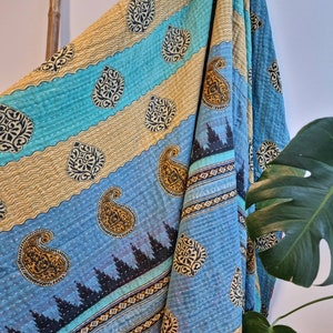 May include: A blue and gold kantha quilt with a paisley pattern. The quilt is folded and draped over a wooden ladder.