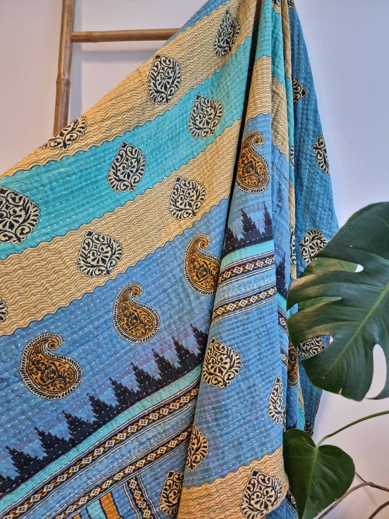 May include: A blue and gold kantha quilt with a paisley pattern. The quilt is folded and draped over a wooden ladder.